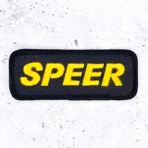 Peut inclure: Un écusson rectangulaire brodé avec un fond noir et une bordure noire épaisse. Le mot SPEER est cousu en lettres capitales jaune vif et audacieuses au centre de l'écusson, avec une police de caractères sans empattement très nette.