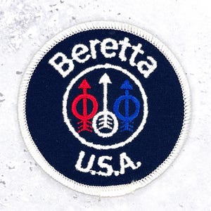 May include: A circular embroidered patch with a dark blue background and a white stitched border. The text Beretta is arched at the top and U.S.A. is at the bottom in white. Three arrows are centred, coloured red, white, and blue respectively.