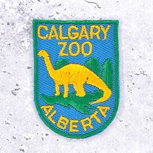 Könnte beinhalten: Gestickter Aufnäher mit einem gelben Dinosaurier auf blauem Hintergrund. Der Text "CALGARY ZOO ALBERTA" ist gelb. Der Aufnäher hat einen grünen Rand. Wahrscheinlich ein Souvenir oder Sammlerstück.