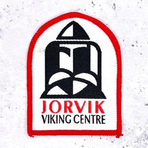 May include: A rectangular patch with a white background, a red border, and a black and white graphic. The patch features the text "JORVIK VIKING CENTRE" in red and black. The graphic is a stylized design.