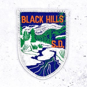 May include: Embroidered patch with a blue background and a white border. The patch features the text "BLACK HILLS S.D." in orange, along with a landscape illustration of mountains, trees, and a river in green and white.