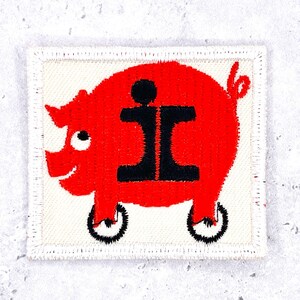 May include: A square embroidered patch featuring a bright red pig on a cream background. The pig has a black letter i on its side and stands on two black wheels. The patch has a stitched border and a curly tail, with a white eye and smile.