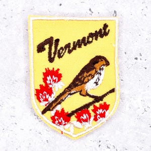 May include: Embroidered patch with a yellow background and a shield shape. The patch features a brown and white bird perched on a branch, red and white flowers, and the word "Vermont" in brown script.