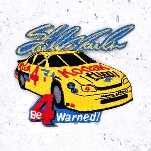 May include: Embroidered patch depicting a yellow race car with the number 4 and the Kodak Film logo. The patch includes the text "Be Warned!" and "Shell/Pennzoil" in blue lettering. The patch is set against a light grey background.