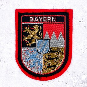 May include: Embroidered patch with a shield design. The patch features the word "BAYERN" at the top. The shield is divided into quadrants with various heraldic symbols in gold, silver, red, and blue. The patch has a red border with black trim.
