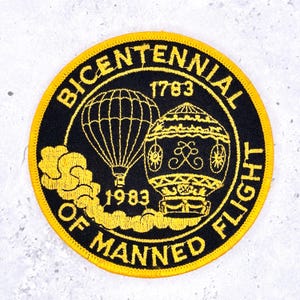 May include: Round black and gold patch commemorating the Bicentennial of Manned Flight. The patch features two hot air balloons, the years 1783 and 1983, and the text "BICENTENNIAL OF MANNED FLIGHT".