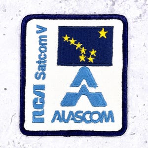 Könnte beinhalten: Quadratischer, gestickter Aufnäher mit weißem Hintergrund, blauem Rand und blauem Text. Der Aufnäher zeigt den Text "RCA Satcom V" und "ALASCOM" sowie ein blaues Rechteck mit gelben Sternen, das einer Flagge ähnelt.