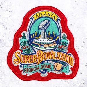 May include: Embroidered patch with a red border featuring the Super Bowl XXVIII logo. The design includes the Atlanta skyline, a football, and the Georgia Dome. The patch has text that reads "Atlanta", "Super Bowl XXVIII", and "Georgia Dome".