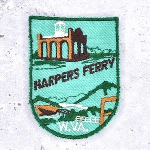 Op de afbeelding: Geborduurde patch met een turquoise achtergrond, een bruin en wit gebouw, groene bergen en de tekst "HARPERS FERRY W.VA." De patch heeft een groene rand en toont een schilderachtig landschap.