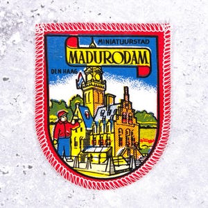 Op de afbeelding: Een schildvormige patch met een rode rand en witte stiksels. De patch heeft een blauwe achtergrond met de tekst "MADURODAM" en "DEN HAAG". Er is ook een kleurrijke illustratie van gebouwen en een persoon aanwezig.