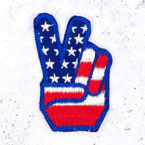 May include: An embroidered patch in the shape of a hand making a peace sign. The index and middle fingers feature a dark blue background with white stars, while the thumb and remaining fingers display horizontal red and white stripes.