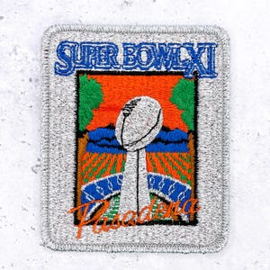 May include: A rectangular embroidered patch featuring a silver border and the blue text SUPER BOWL XI at the top. The center depicts a white football trophy against an orange and green landscape with blue accents and the orange text Pasadena below.