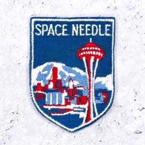 May include: Embroidered patch featuring the Space Needle in Seattle. The patch has a blue background with white text that reads "SPACE NEEDLE." The design includes a red and white Space Needle, a mountain range, and city buildings.