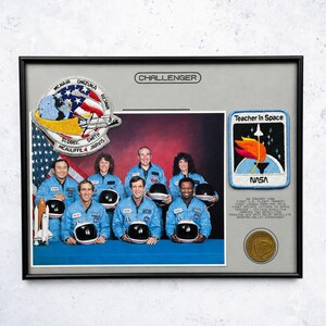 May include: A framed commemorative display featuring the Challenger crew in blue flight suits, with a mission patch, a Teacher in Space patch, a small space shuttle model, and a gold coin. Text includes Challenger, Teacher in Space, and mission details.