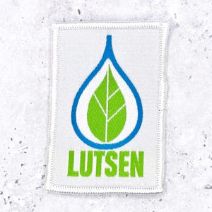 Op de afbeelding: Een rechthoekige geborduurde patch met een witte achtergrond en een gestikte rand. Het ontwerp toont een blauwe waterdruppelomtrek met daarin een felgroen blad met witte nerven. Onder de afbeelding is het woord LUTSEN in het groen geborduurd.