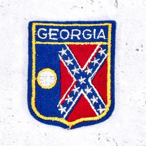 以下が含まれることがあります： 上部に「GEORGIA」の文字が入った刺繍パッチ。パッチは青い縁取りと黄色の輪郭があります。デザインには、白い星が付いた赤い十字、青いフィールド、白い円が含まれています。