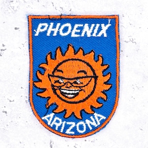 May include: A rectangular embroidered patch with a blue background and an orange border. The patch features a smiling sun character wearing glasses in the center, with the white text PHOENIX at the top and the white text ARIZONA at the bottom.