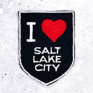 以下が含まれることがあります： 白い縁取りのある黒いパッチ。白い文字で「I」と「SALT LAKE CITY」の文字が書かれており、その間に赤いハートがあります。パッチは盾の形をしています。