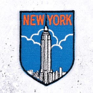 Może przedstawiać: Naszywka w kształcie tarczy z niebieskim tłem, przedstawiająca biały zarys Empire State Building i otaczających go wieżowców. Napis NEW YORK jest wyhaftowany na górze w kolorze pomarańczowym, z białymi chmurami za budynkiem.