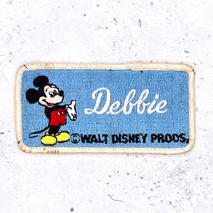 May include: Rectangular patch with a light blue background featuring Mickey Mouse and the name "Debbie" in white script. The patch has a yellow border and the text "WALT DISNEY PRODS." at the bottom.