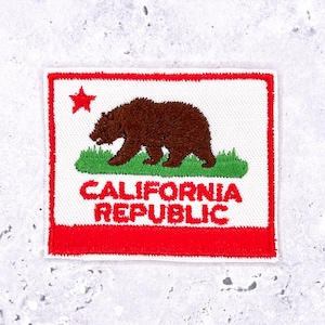 May include: Embroidered patch featuring the California state flag design. The patch has a white background with a brown bear, green grass, and a red star in the upper left corner. The words "CALIFORNIA REPUBLIC" are in red.