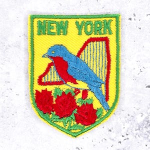 May include: Embroidered patch featuring a blue bird with red accents perched above red roses and a harp, set against a yellow background. The patch is bordered in green and has the words "NEW YORK" embroidered in green.