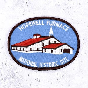 Op de afbeelding: Ovale patch met een gebouw met een rood dak en witte muren op een lichtblauwe achtergrond. De tekst "HOPEWELL FURNACE NATIONAL HISTORIC SITE" is in het wit geborduurd, met een bruine rand. Afbeelding van een historische plek.
