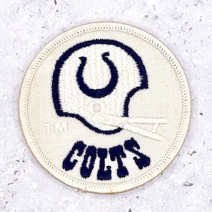 May include: A circular patch featuring the Indianapolis Colts logo. The design includes a blue horseshoe inside a football helmet outline, with the word "COLTS" in blue below. The patch has a cream-colored background and a stitched border.