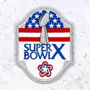 Op de afbeelding: Geborduurde patch met het Super Bowl X logo. Het ontwerp bevat een zilveren voetbaltrofee, rood-witte strepen, blauwe sterren en de woorden "SUPER BOWL X" in blauw. De patch heeft een zilveren rand.