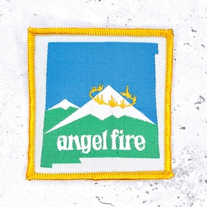 Op de afbeelding: Vierkantige patch met een gele geborduurde rand. De patch heeft een blauwe lucht, witte bergen en groene heuvels. De tekst "angel fire" is in het wit en een gouden ontwerp staat bovenop de berg.
