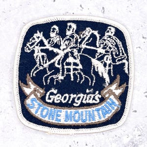 May include: A navy blue embroidered patch featuring a white depiction of three figures on horseback. The patch has a white border and includes the text "Georgia's STONE MOUNTAIN" in a banner.