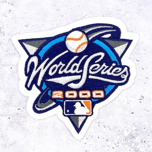May include: Embroidered patch featuring the words "World Series 2000" in white script. The patch has a baseball and the MLB logo. The design includes blue, orange, gray, and white colors. The patch is in the shape of a triangle.
