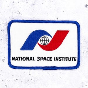 Pode incluir: Um emblema retangular bordado com fundo branco e borda azul. Apresenta uma forma curva azul, uma forma curva vermelha e um pequeno ícone de globo preto no centro. O texto NATIONAL SPACE INSTITUTE está bordado em preto.