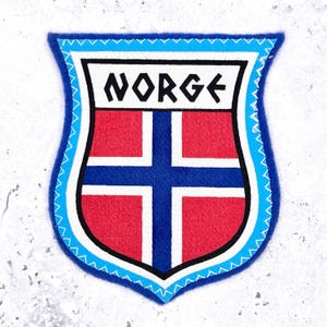 May include: A shield-shaped embroidered patch featuring the word NORGE in black lettering on a white background at the top. Below, a red field is divided by a blue cross with white borders. The entire emblem is outlined in bright blue zigzag stitching.