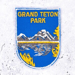 May include: A shield-shaped embroidered patch featuring a blue background with the yellow text GRAND TETON PARK at the top. The design depicts grey and white snow-capped mountains above a grey lake reflection, with yellow trees on the sides.