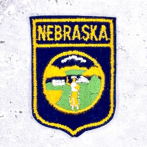 May include: Embroidered patch featuring the word "NEBRASKA" in yellow, above a circular design with a golfer. The patch has a navy blue background with a yellow border. The golfer is in a yellow outfit, with green and white details.