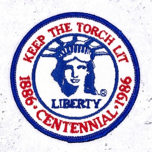 May include: A round patch with a blue border and the text "KEEP THE TORCH LIT" in red. The center features a blue image of the Statue of Liberty and the word "LIBERTY". The text "1886 CENTENNIAL 1986" is also included.