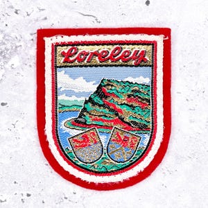 May include: A Loreley patch with a red border and a white edge. The patch features a landscape scene with the word "Loreley" at the top. The scene includes a mountain, water, and two crests at the bottom.