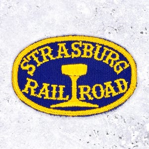 Könnte beinhalten: Gestickter Aufnäher mit blauem ovalem Hintergrund und gelbem Text und Rand. Der Text lautet "STRASBURG RAIL ROAD" mit einer gelben stilisierten Zugwagengrafik in der Mitte. Der Aufnäher ist wahrscheinlich für Eisenbahnliebhaber.