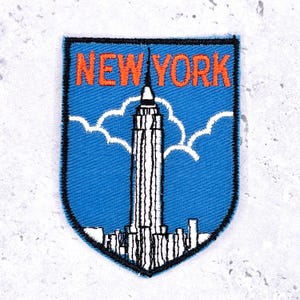 May include: Embroidered patch featuring the words "NEW YORK" in orange, above a black and white depiction of the Empire State Building. The patch has a blue background with white cloud-like shapes and a black border.