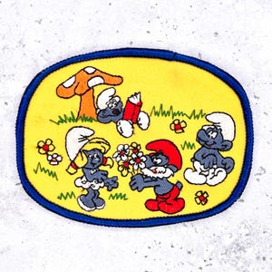 May include: An oval-shaped patch featuring a scene with Smurfs. The patch has a yellow background with a blue border. The Smurfs are in various poses, with a mushroom and flowers in the scene. The Smurfs are wearing white hats and pants.
