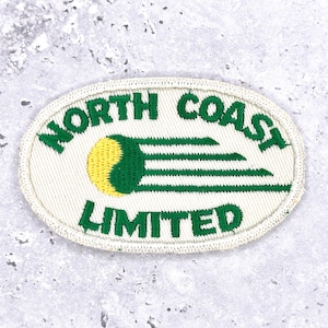 Könnte beinhalten: Ein ovales Abzeichen mit dem grünen Schriftzug "NORTH COAST LIMITED". Das Design zeigt ein gelb-grünes Emblem mit grünen horizontalen Linien. Das Abzeichen ist mit weißem Stoff eingefasst.