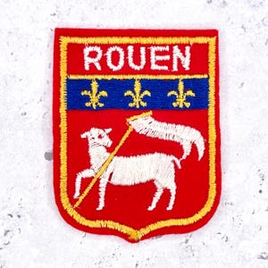 Peut inclure: Un écusson brodé rectangulaire à fond rouge avec une bordure jaune. La partie supérieure affiche le texte blanc ROUEN. En dessous, une bande horizontale bleue porte trois fleurs de lys jaunes. Un agneau blanc tient un drapeau blanc sur une hampe.