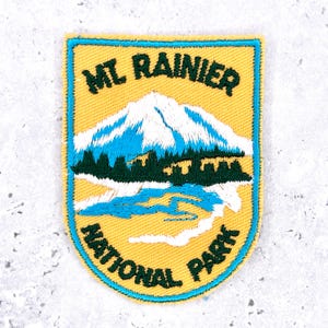 May include: A shield-shaped embroidered patch with a yellow background and a bright blue border. It features a white snow-capped mountain, dark green pine trees, and a blue river. The text reads MT RAINIER at the top and NATIONAL PARK at the bottom.