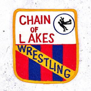 May include: A rectangular embroidered patch with a yellow border. The top half is white with red text reading CHAIN OF LAKES and a circular icon of two wrestlers. The bottom half features vertical red and blue stripes with a yellow diagonal banner.