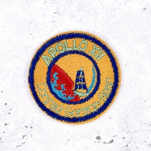 Pode incluir: Um emblema bordado circular com fundo dourado e borda azul escura. O texto APOLLO XII está bordado em verde claro no topo, com Conrad Gordon Bean na parte inferior. Um gráfico vermelho e azul retrata um módulo espacial.