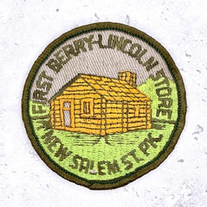 Op de afbeelding: Ronde geborduurde patch met een geel blokhutontwerp, tegen een groene achtergrond en boom. De patch bevat de tekst "FIRST BERRY-LINCOLN STORE NEW SALEM ST. PA" in groen en geel, met een donkergroene rand.