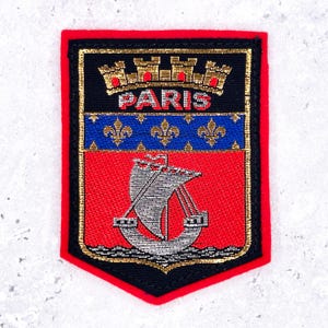 May include: A rectangular embroidered patch featuring the coat of arms of Paris. The design includes a gold mural crown above the word PARIS, a blue band with three gold fleur-de-lis, and a silver ship on a red field, all bordered in red.