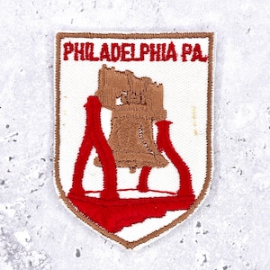 Könnte beinhalten: Gestickter Aufnäher mit der Freiheitsglocke und dem Text "PHILADELPHIA PA" in roter Farbe auf weißem Hintergrund. Der Aufnäher hat einen braunen Rand und ist schildförmig.