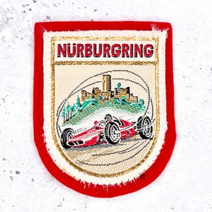 May include: A rectangular embroidered patch with a red border and a cream-colored center. The text NÜRBURGRING is stitched in red at the top. Below, a red race car is depicted driving on a track in front of a castle and green trees.